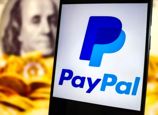 PayPal logo on a phone in front of a defocused background of gold coins and Benjamin Franklin's portrait from the US $100 bill